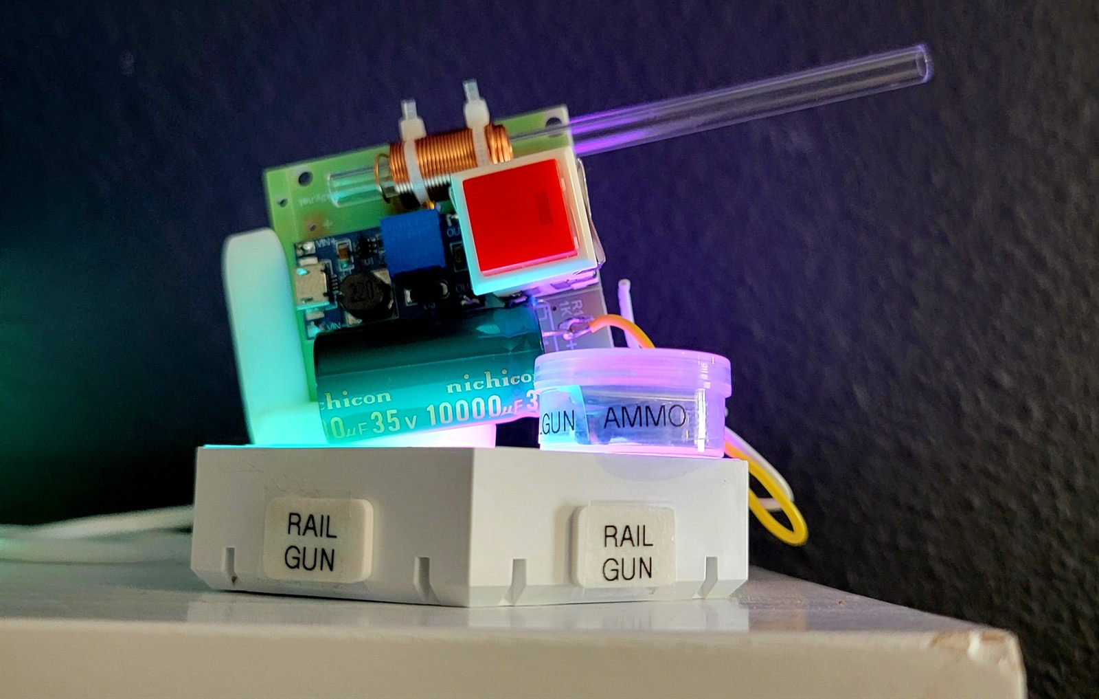 Small first-build launcher on a white 3D-printed base: a coil and driver PCB, two 10,000 microfarad capacitors, a red illuminated fire button, a clear acrylic barrel and keycaps labelled RAIL GUN and AMMO, lit by colour-changing LEDs.