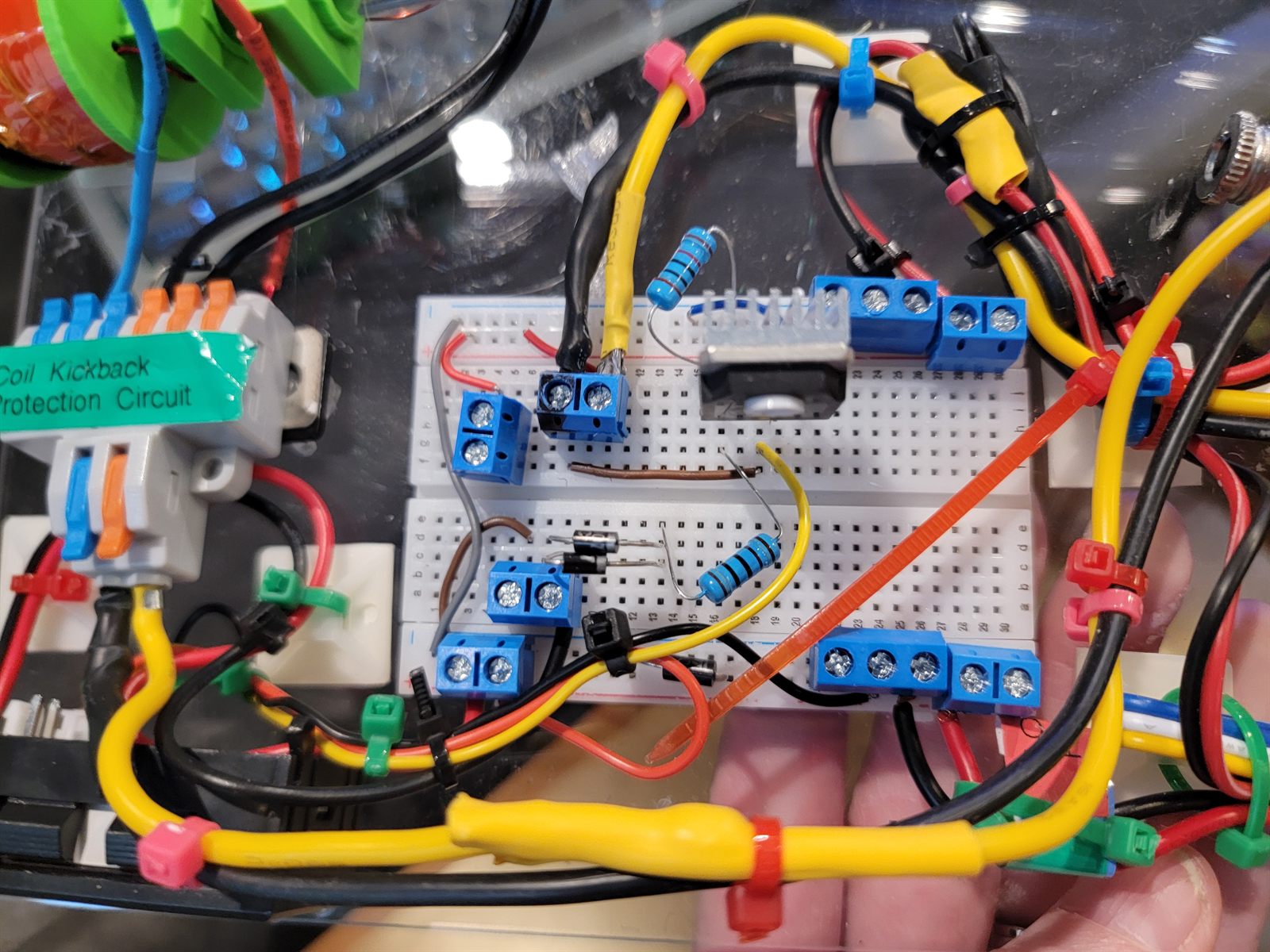 Hand holding a solderless breadboard carrying the coil kickback protection circuit — diode, resistors and terminal blocks — surrounded by yellow and red silicone wire and lever-nut connectors, with a green label reading Coil Kickback Protection Circuit.