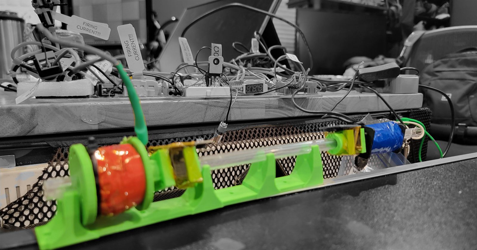 Green 3D-printed rail holding a potted red coil, a clear barrel tube and a capacitor, on a bench where several cables carry paper flags reading FIRING CURRENT.