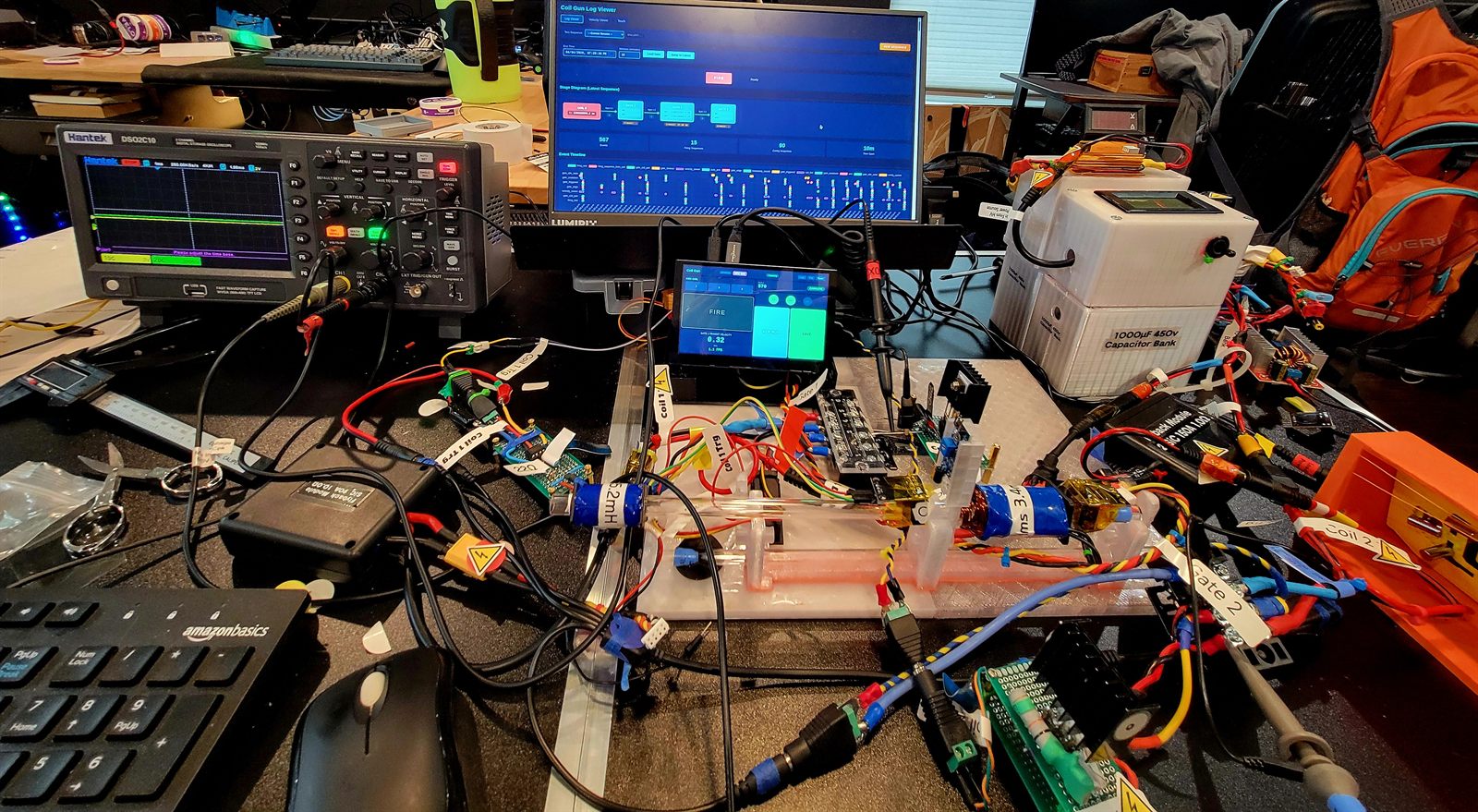 Workbench with an oscilloscope, a log-viewer monitor, a 450 volt capacitor bank and a three-coil launcher rail wired up under scope probes.