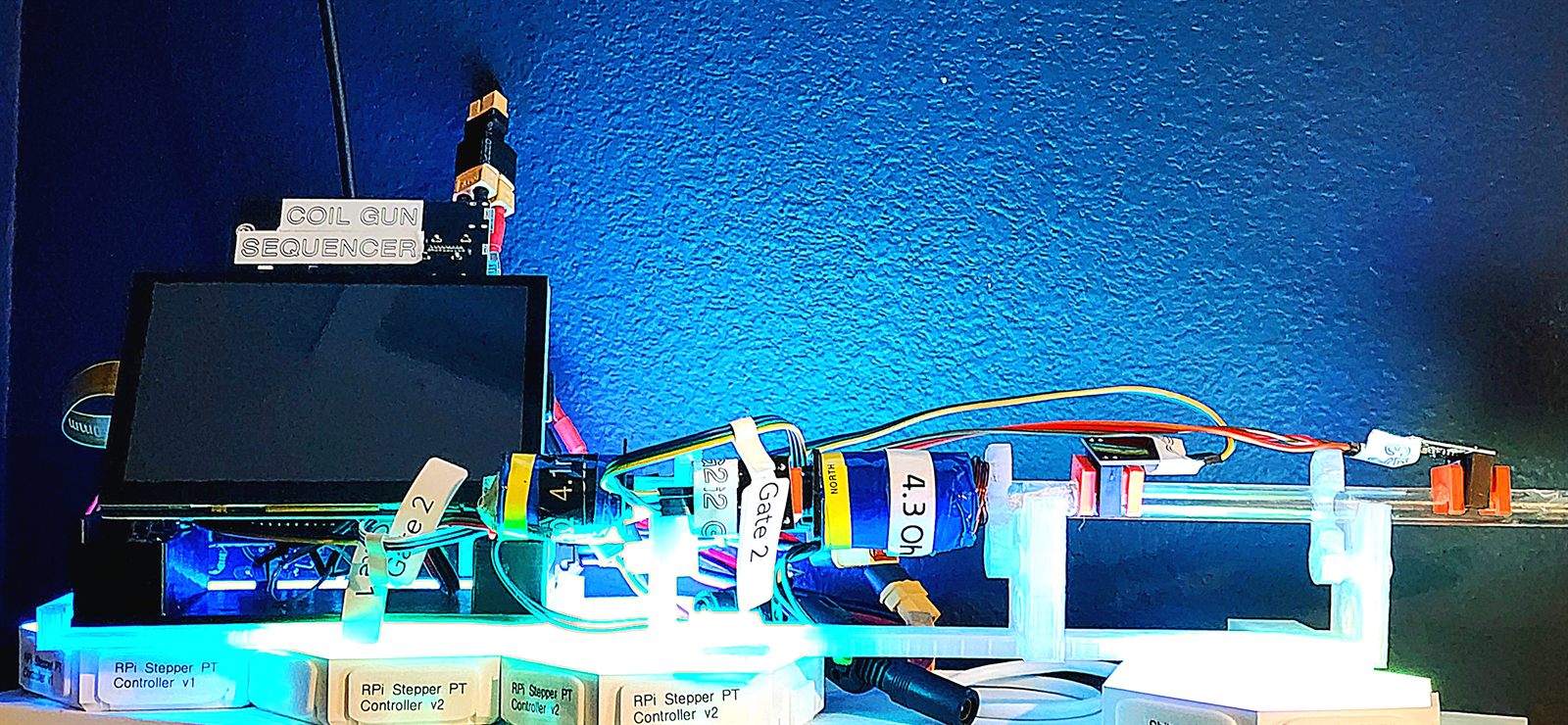 Night bench under blue light: the sequencer enclosure and touchscreen beside the launcher rail, with a coil labelled 4.3 ohm and NORTH, gate flags, and a row of printed pedestals labelled RPi Stepper PT Controller v1 and v2.