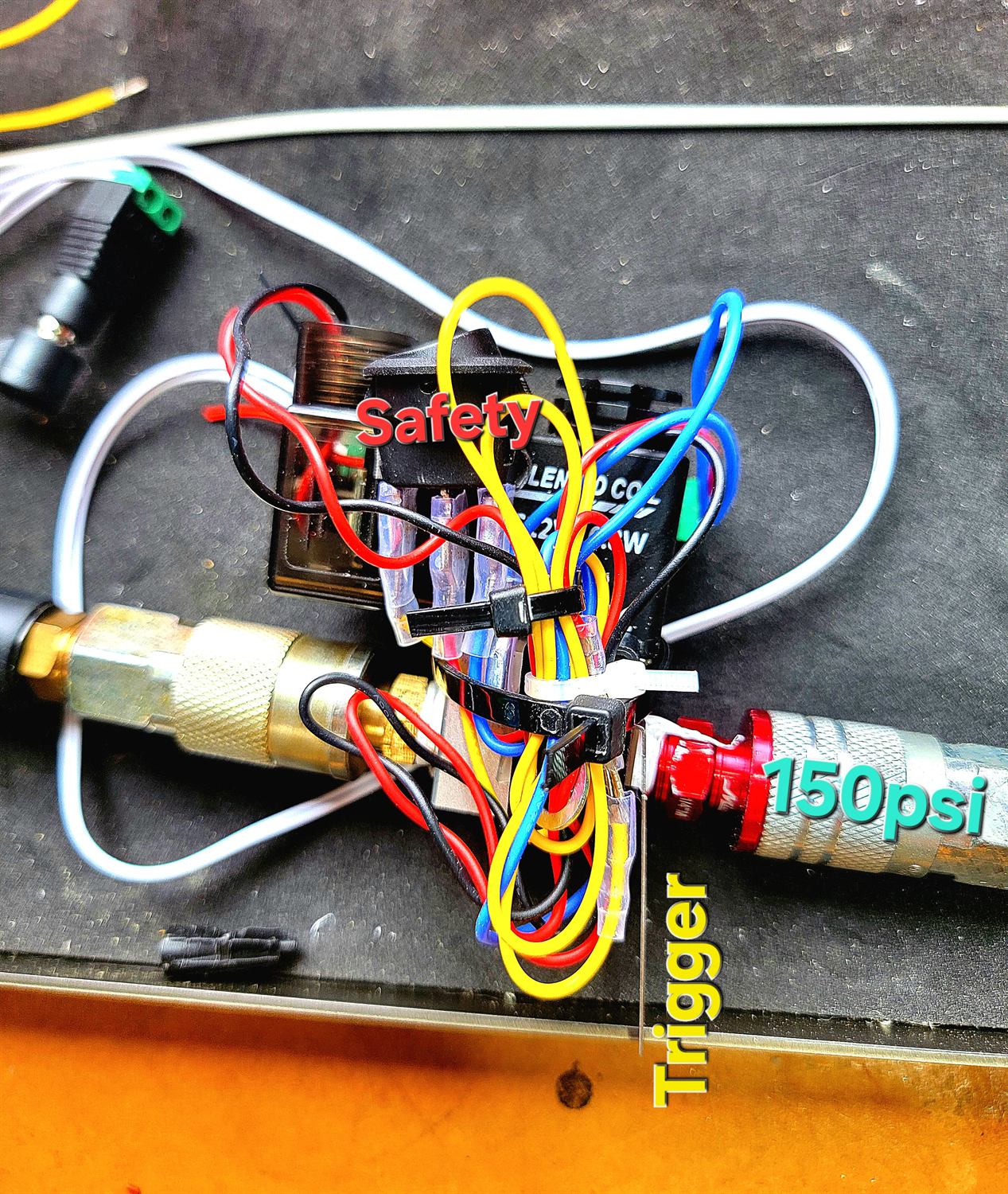 Overhead photo of a 24 volt solenoid valve wired into brass quick-connect air fittings, hand-annotated with the words Safety, Trigger and 150psi pointing at the relay, the switch line and the red regulator fitting.