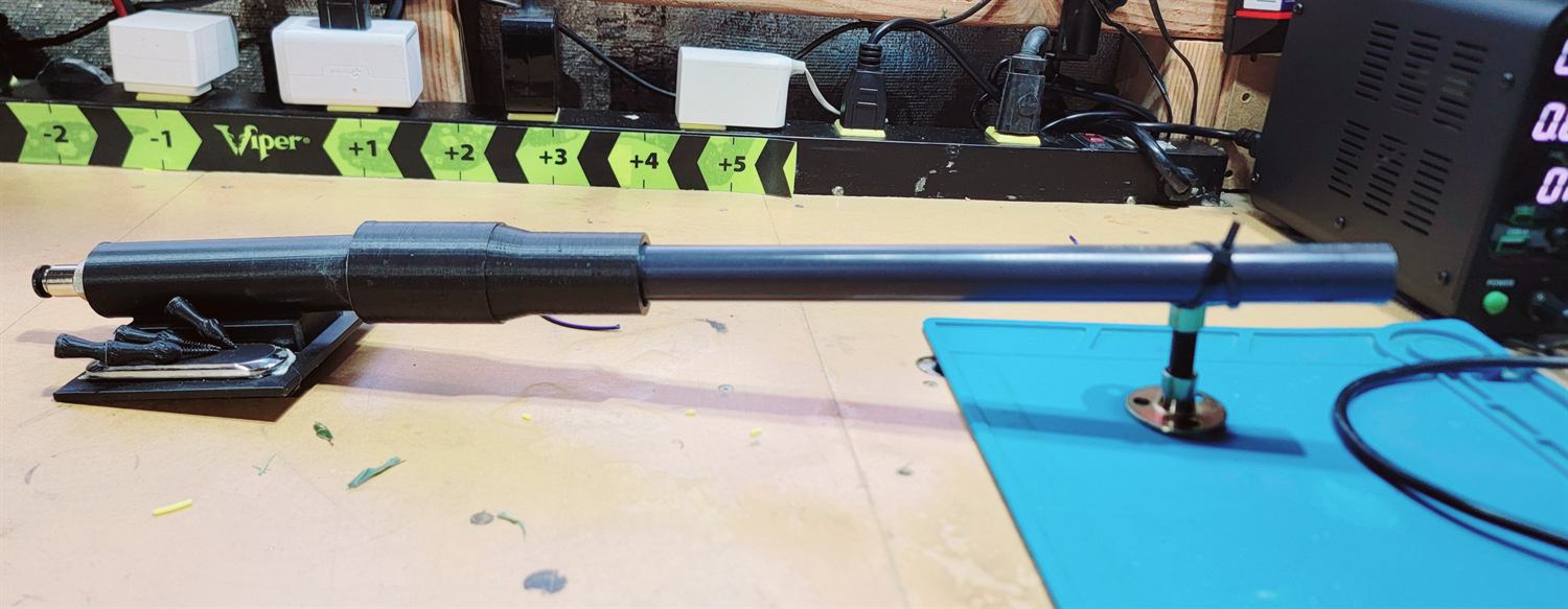 Black pressure chamber and steel barrel assembled on a low stand on a workbench, next to a bench power supply.