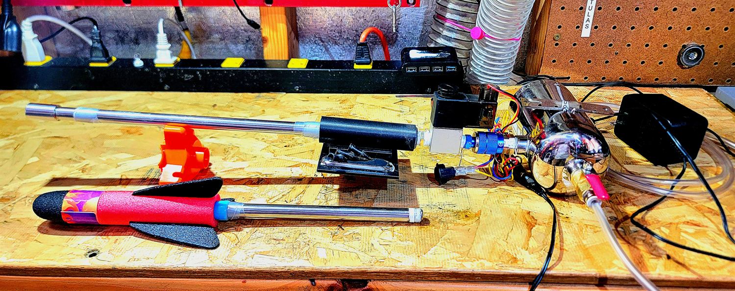 Workbench with a foam-tipped toy rocket beside two candidate barrels, a red printed cradle, and the solenoid valve assembly plumbed to a polished air chamber.