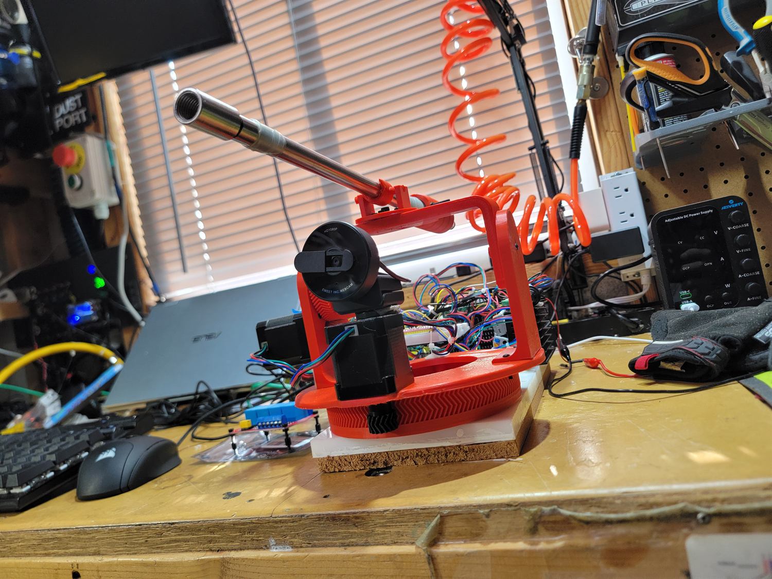 Red 3D-printed pan and tilt yoke on a large printed turntable, carrying a webcam and a steel barrel, mounted on a cork board with an orange coiled air hose behind it.