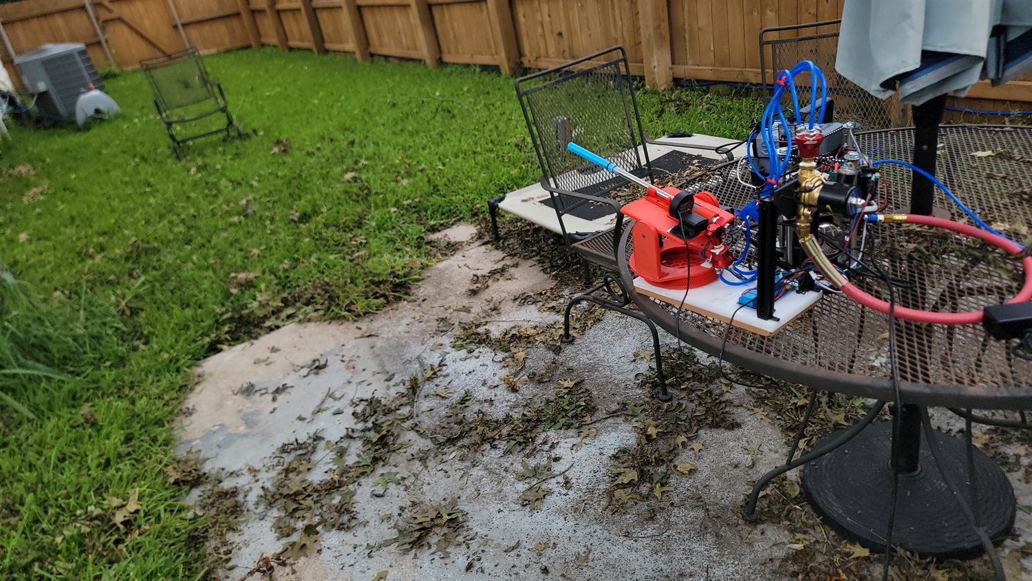 The turret rig set up on a mesh patio table in a fenced back garden, plumbed with blue tubing and a red air hose, aimed across the lawn.