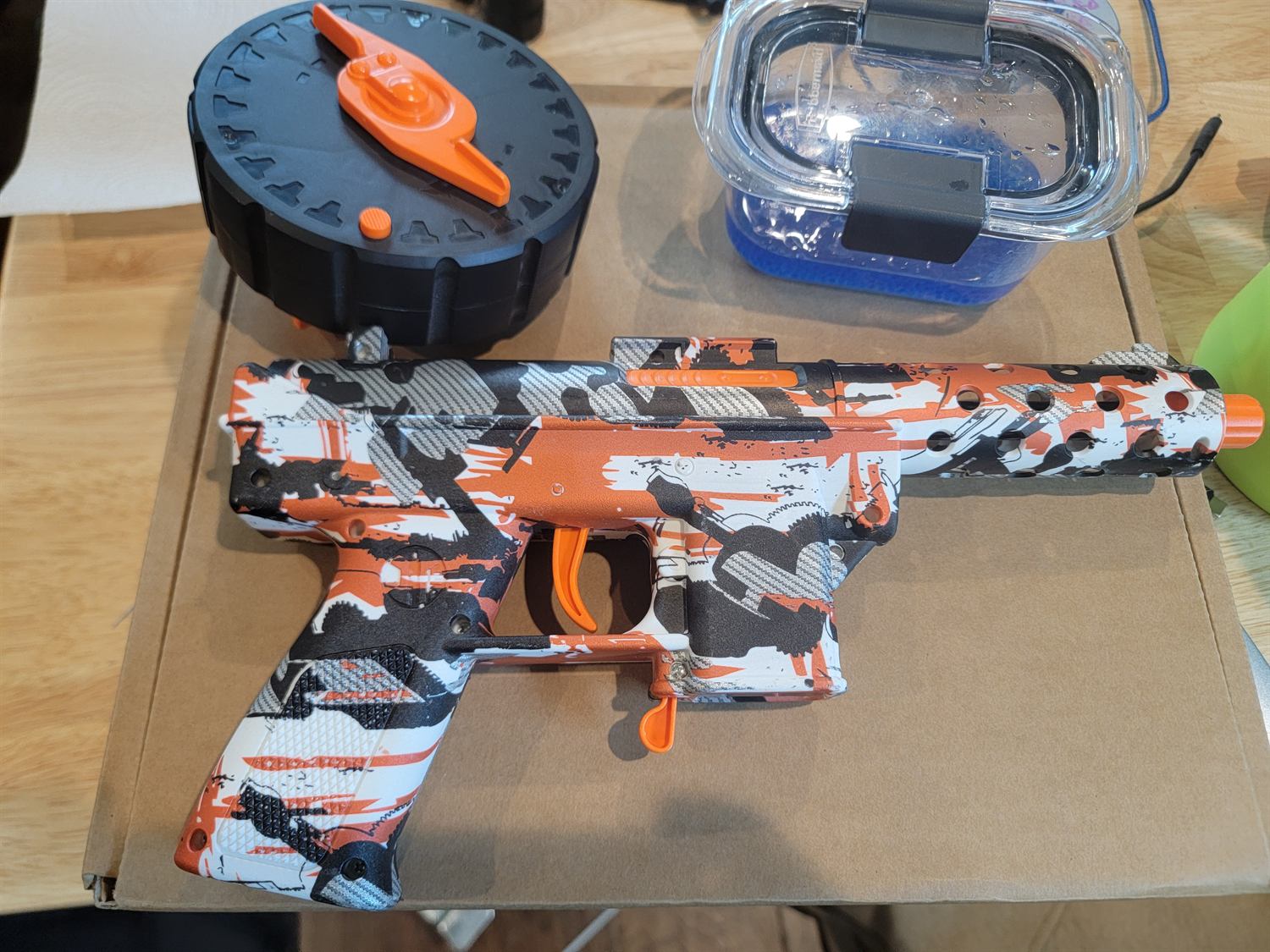 An orange and white camouflaged gel blaster on cardboard beside its black drum magazine and a tub of blue hydrated gel beads.