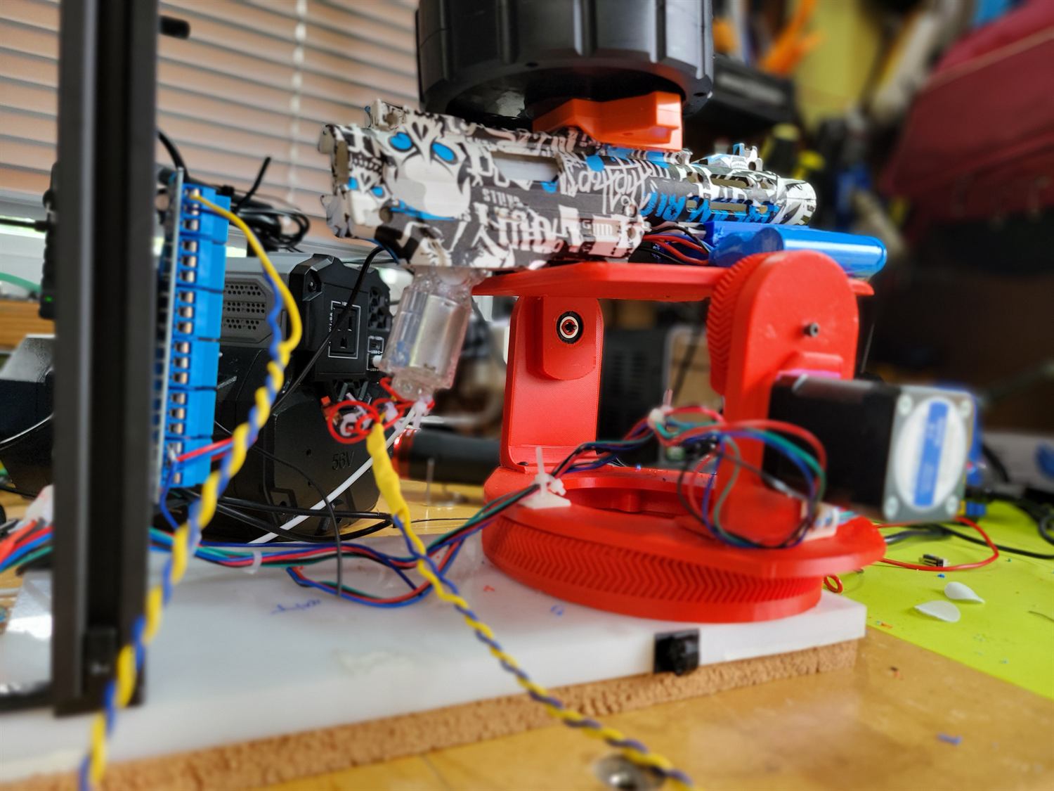 The red turret yoke carrying a graffiti-patterned gel blaster with drum magazine and a clear bead feeder, wired to a blue terminal breakout board.