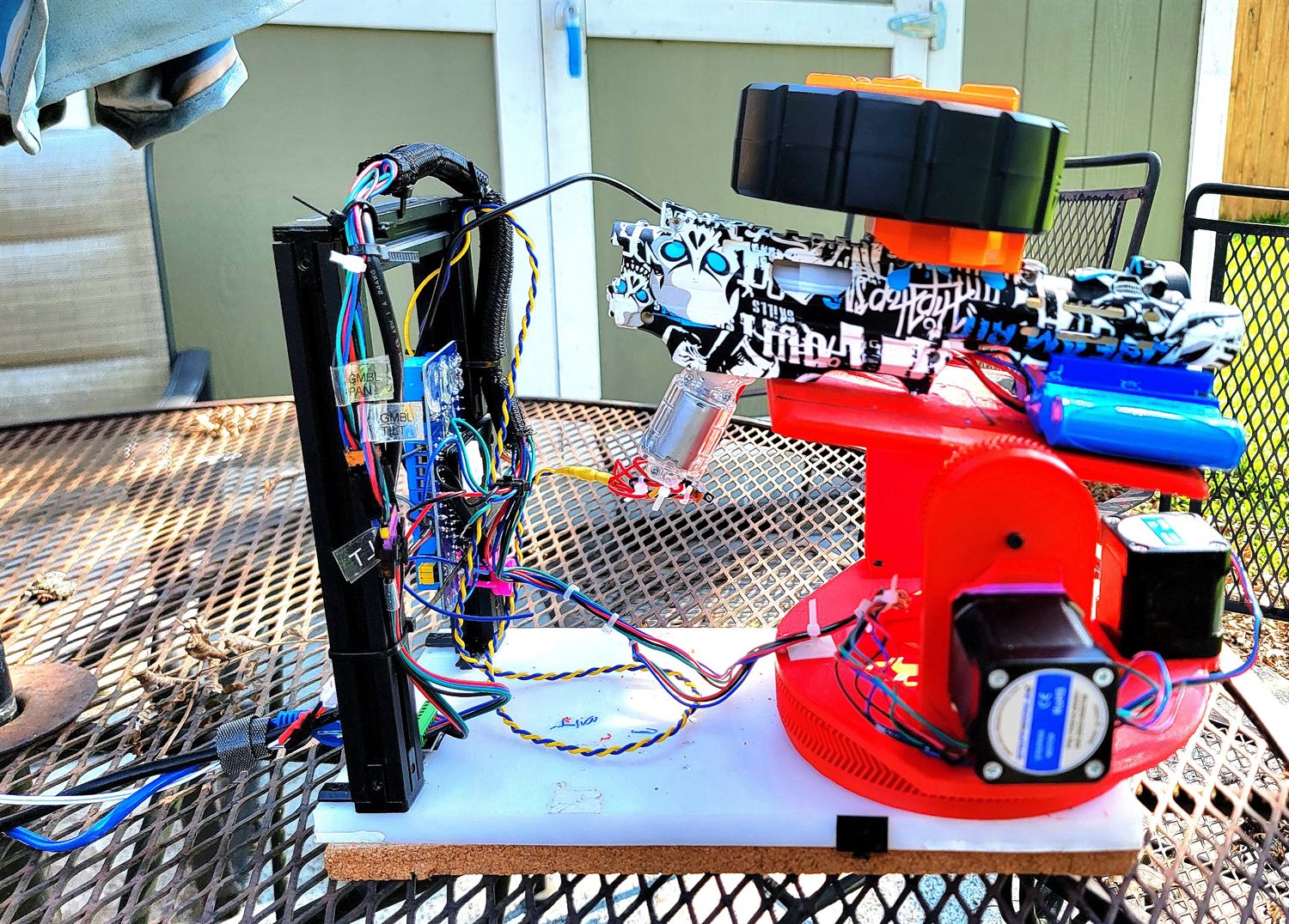 Red 3D-printed pan-tilt turret on a patio table carrying a gel blaster with a drum magazine, wired to a labelled stepper-driver frame.