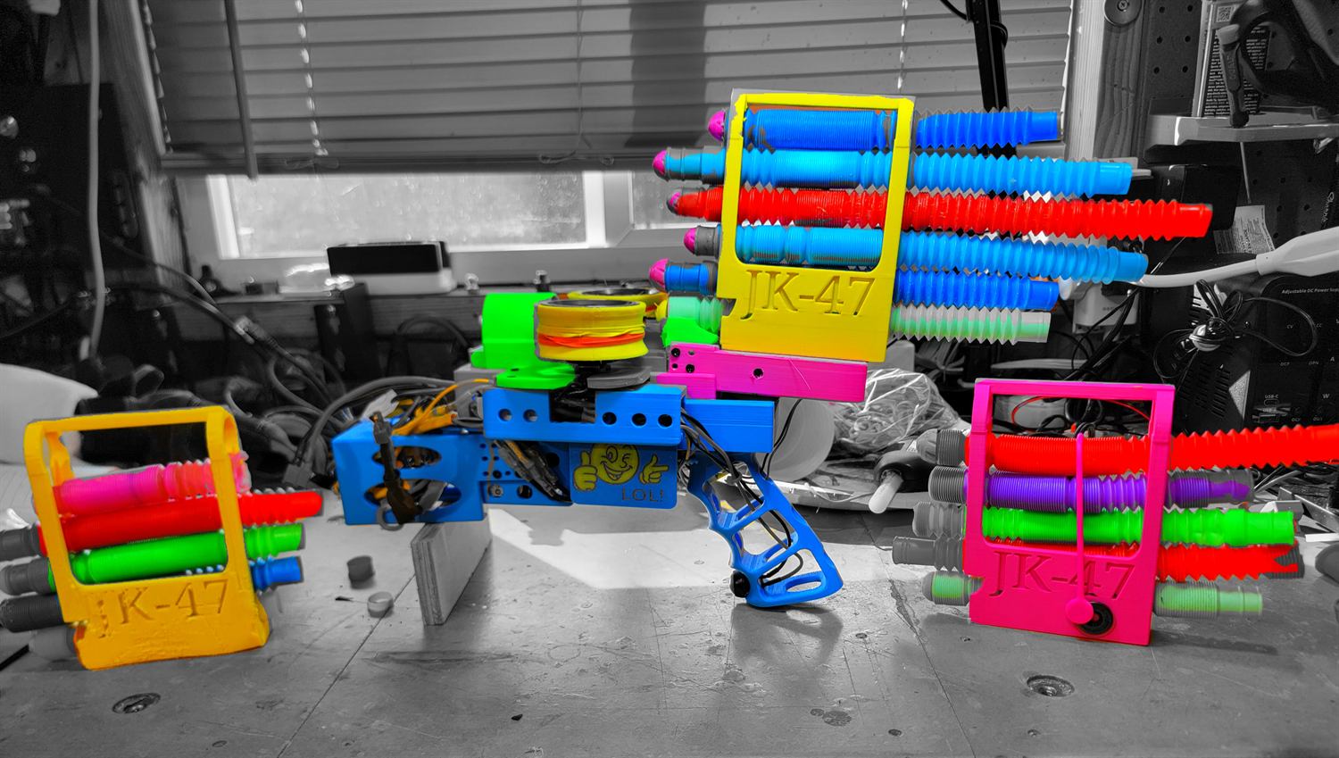 Brightly coloured 3D-printed magazine frames stamped JK-47, each loaded with foam pool-noodle segments, flanking a printed launcher with a blue pistol grip.