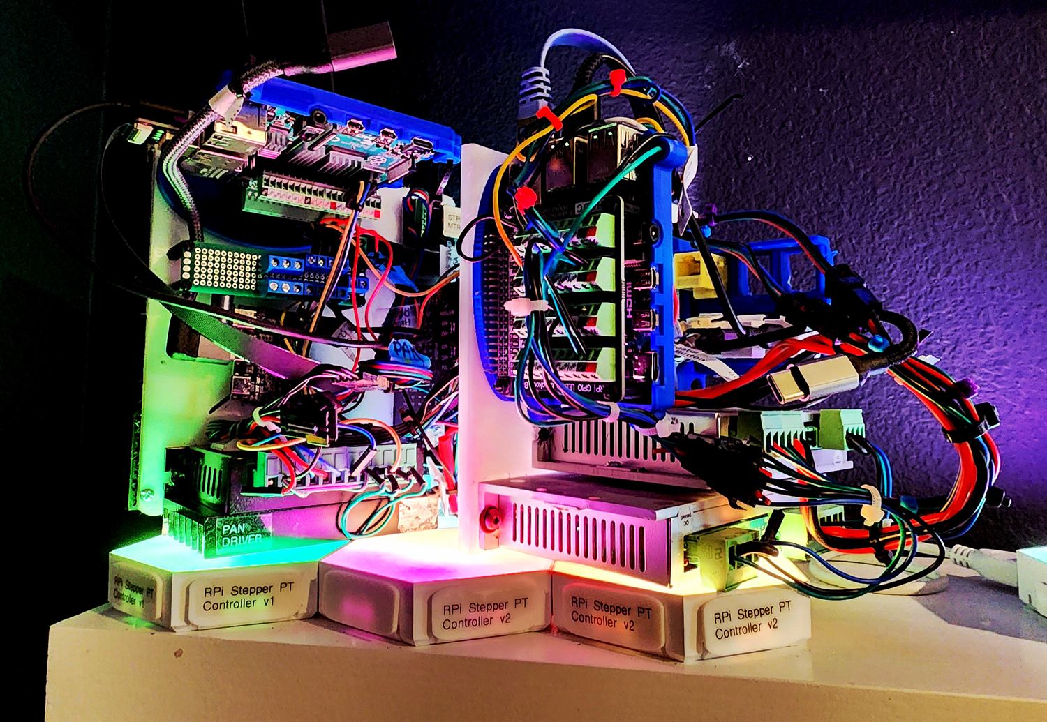 RGB-lit shelf of Raspberry Pi stepper controller assemblies standing on printed pedestals labelled RPi Stepper PT Controller v1 and three labelled v2, with one board marked PAN DRIVER.